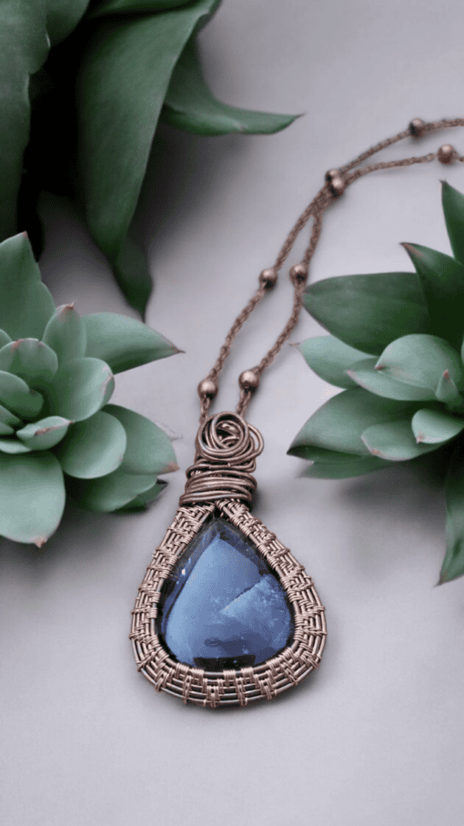 A wire-wrapped necklace with a teardrop-shaped blue gemstone pendant, placed between two green succulents.