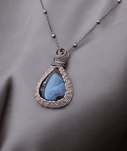 Bronze wire-wrapped pendant with a blue teardrop gemstone on a delicate chain with evenly spaced beads, laid on gray fabric.