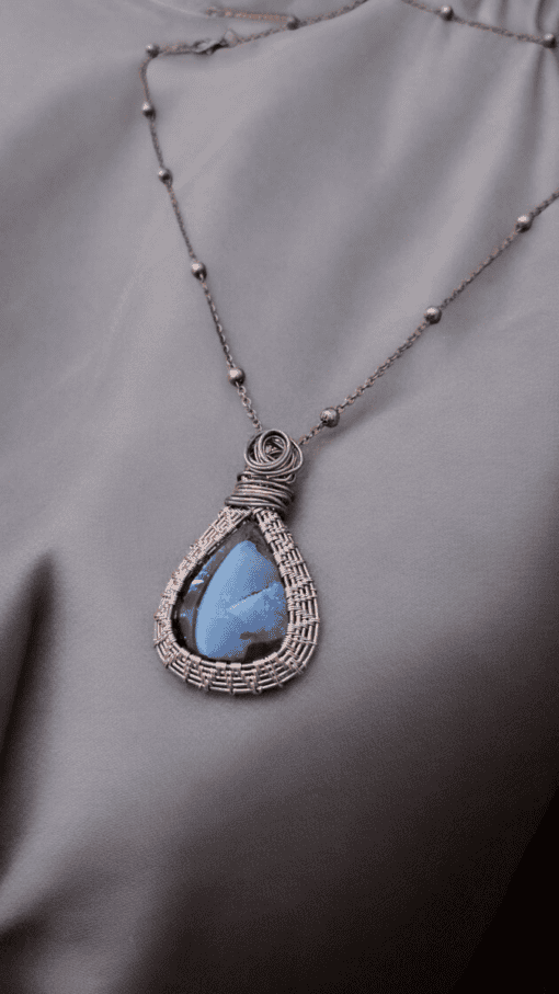 Bronze wire-wrapped pendant with a blue teardrop gemstone on a delicate chain with evenly spaced beads, laid on gray fabric.