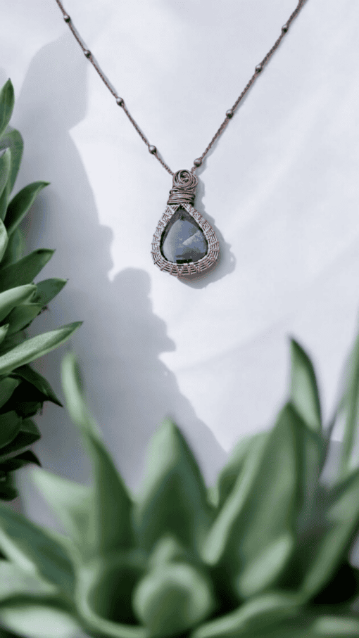 Bronze wire-wrapped necklace with a teardrop-shaped blue stone, framed by green succulent plants.