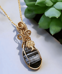 Gold wire-wrapped pendant with a striped black stone and tree design, displayed against a blurred background with green succulents.