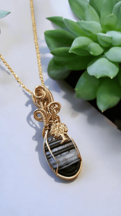 Gold wire-wrapped pendant with a striped black stone and tree design, displayed against a blurred background with green succulents.