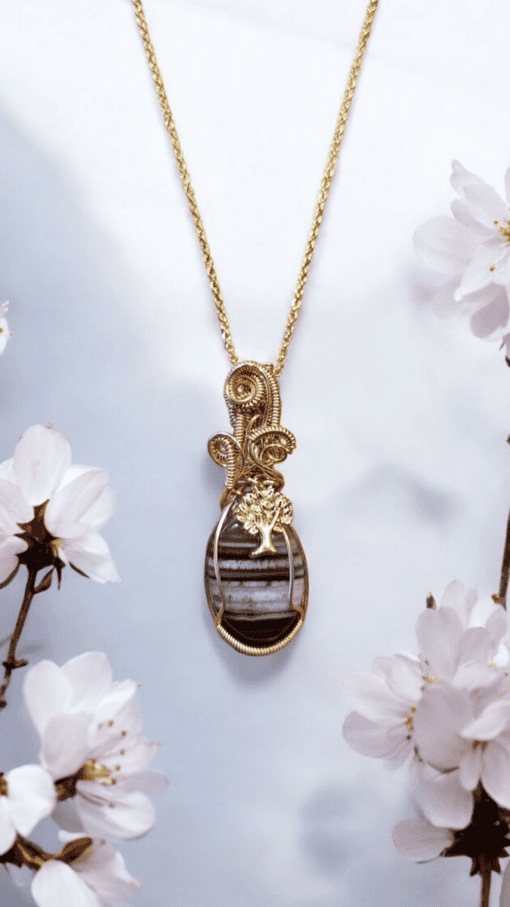 Gold necklace with an intricate pendant featuring a tree design, surrounded by white flowers on a light background.