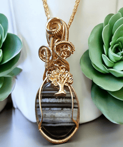 Gold wire-wrapped pendant with a striped stone and tree charm, displayed between two green succulent plants.