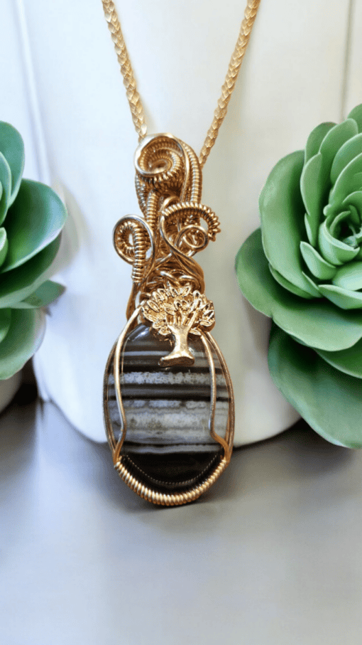 Gold wire-wrapped pendant with a striped stone and tree charm, displayed between two green succulent plants.