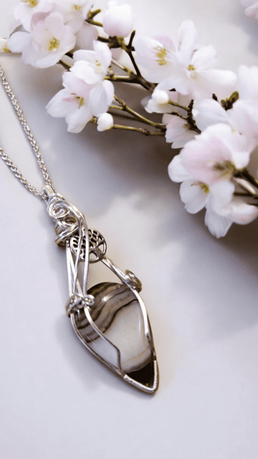 Silver wire-wrapped pendant with a striped stone, set against white and pink cherry blossom flowers on a light background.