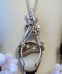 Wire-wrapped stone pendant with intricate silver design, featuring a tree of life symbol, surrounded by blurred pink flowers.