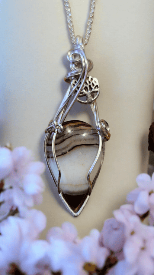 Wire-wrapped stone pendant with intricate silver design, featuring a tree of life symbol, surrounded by blurred pink flowers.