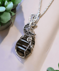 Silver necklace with a striped pendant, featuring intricate wire designs and a moon motif, surrounded by green succulent plants.