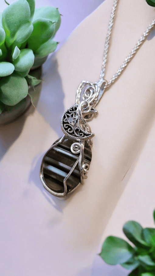 Silver necklace with a striped pendant, featuring intricate wire designs and a moon motif, surrounded by green succulent plants.