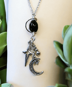 Silver necklace with a hanging black bead, star, crescent moon, and small spheres, surrounded by green leaves.