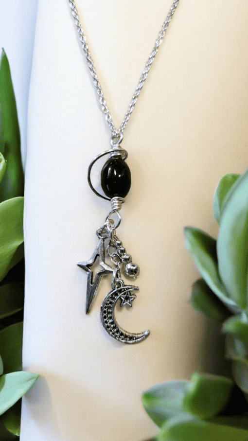 Silver necklace with a hanging black bead, star, crescent moon, and small spheres, surrounded by green leaves.