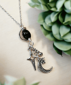 Silver necklace with star and crescent moon charms on a light background, surrounded by green succulent plants.