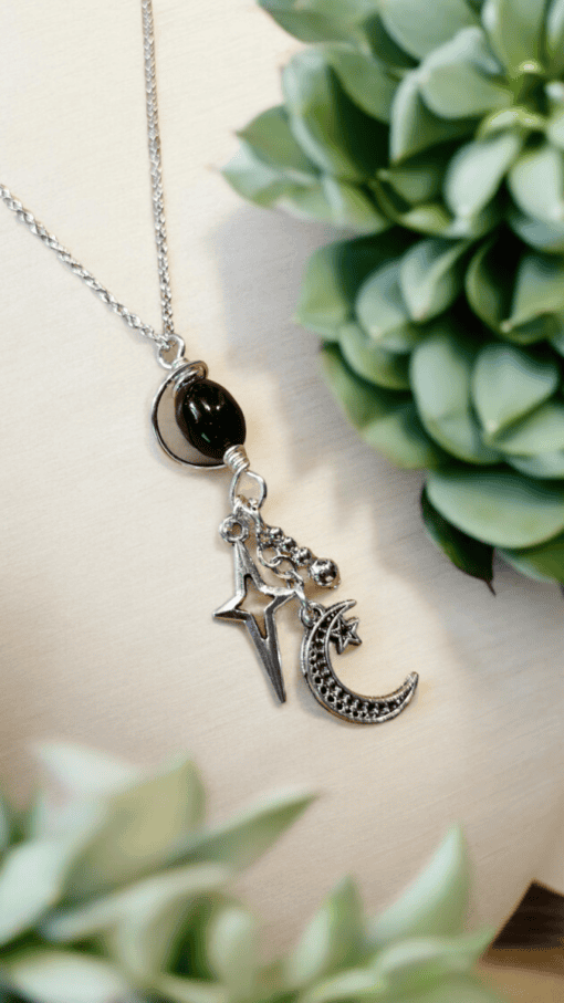 Silver necklace with star and crescent moon charms on a light background, surrounded by green succulent plants.