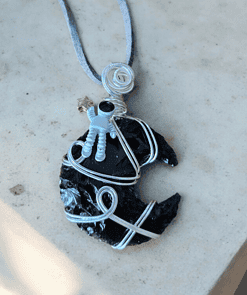 A necklace with a pendant featuring a small astronaut figure on a wire-wrapped black stone, resembling the shape of a crescent moon.