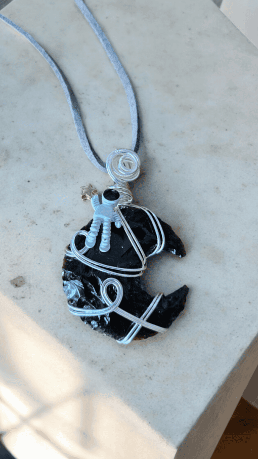 A necklace with a pendant featuring a small astronaut figure on a wire-wrapped black stone, resembling the shape of a crescent moon.