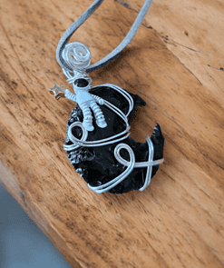 A polymer clay astronaut holding a star sits on a wire-wrapped black crescent moon pendant with a grey cord necklace.