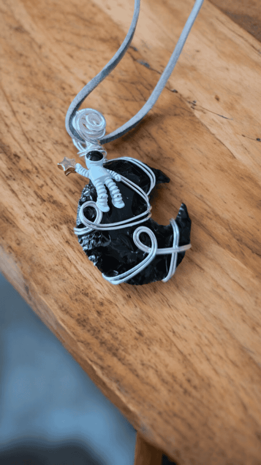 A polymer clay astronaut holding a star sits on a wire-wrapped black crescent moon pendant with a grey cord necklace.