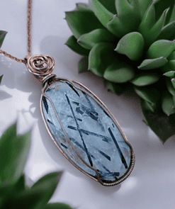 A wire-wrapped blue gemstone pendant on a chain, surrounded by green succulent plants.