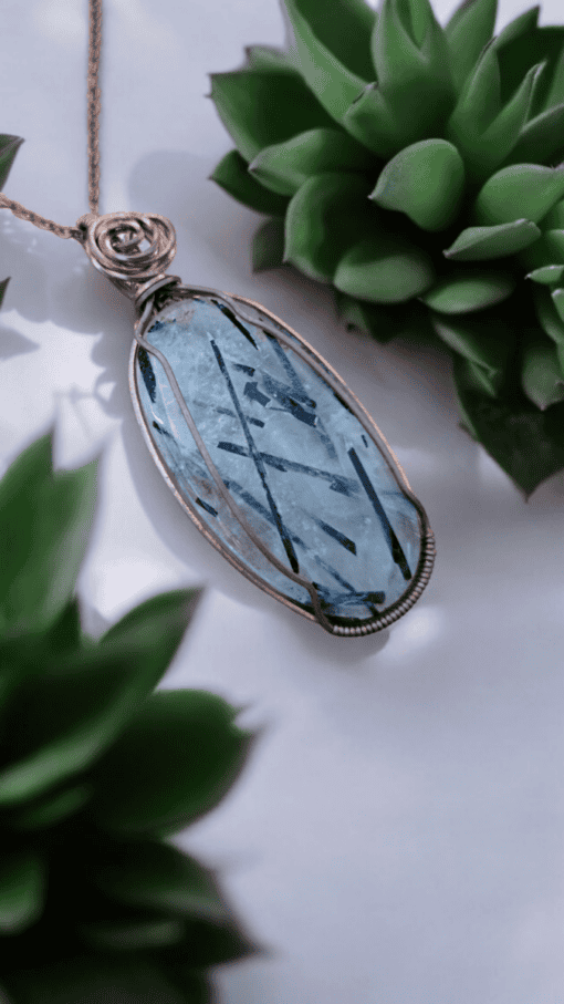 A wire-wrapped blue gemstone pendant on a chain, surrounded by green succulent plants.