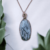 Pendant with a blue gemstone and intricate wire wrapping, displayed against a white background with green succulent plants.