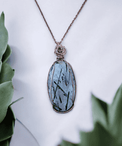 Pendant with a blue gemstone and intricate wire wrapping, displayed against a white background with green succulent plants.