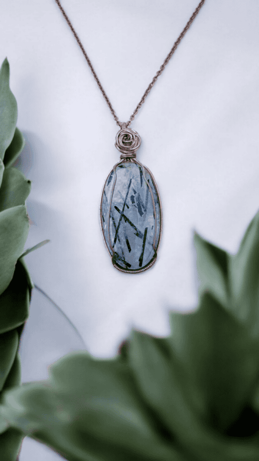 Pendant with a blue gemstone and intricate wire wrapping, displayed against a white background with green succulent plants.