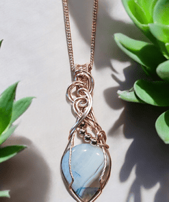 Gold wire-wrapped pendant with blue stone on a chain, bordered by green succulent plants on a light background.