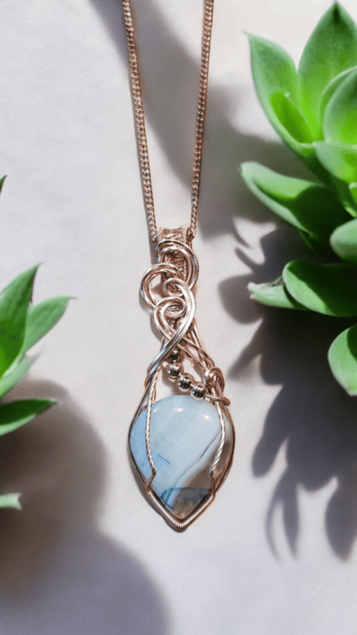 Gold wire-wrapped pendant with blue stone on a chain, bordered by green succulent plants on a light background.