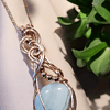 A wire-wrapped pendant with a blue gemstone and metallic accents hangs from a chain, placed next to a green succulent plant.