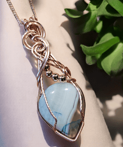 A wire-wrapped pendant with a blue gemstone and metallic accents hangs from a chain, placed next to a green succulent plant.