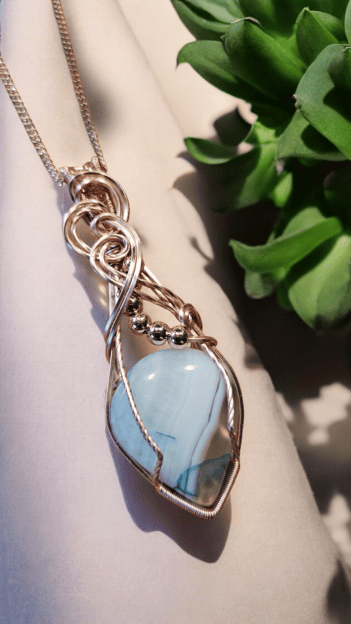 A wire-wrapped pendant with a blue gemstone and metallic accents hangs from a chain, placed next to a green succulent plant.