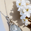 A teardrop-shaped blue agate pendant with intricate silver wire wrapping, resting beside white flowers on a beige background.