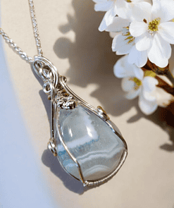 A teardrop-shaped blue agate pendant with intricate silver wire wrapping, resting beside white flowers on a beige background.