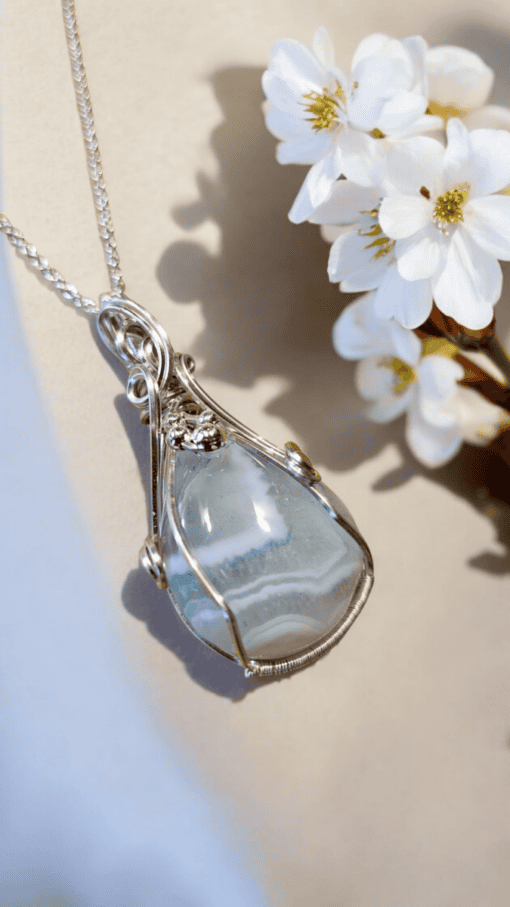 A teardrop-shaped blue agate pendant with intricate silver wire wrapping, resting beside white flowers on a beige background.