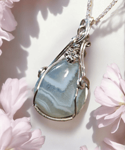 Silver pendant with a blue agate stone and small flower embellishments, surrounded by soft pink flower petals.