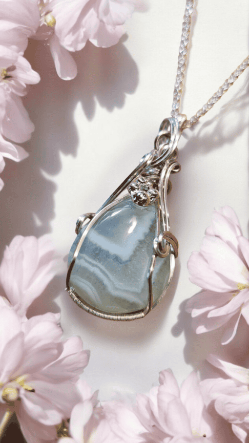 Silver pendant with a blue agate stone and small flower embellishments, surrounded by soft pink flower petals.