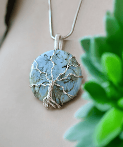 Silver wire-wrapped blue stone necklace with a tree of life design, displayed near a green plant on a beige surface.