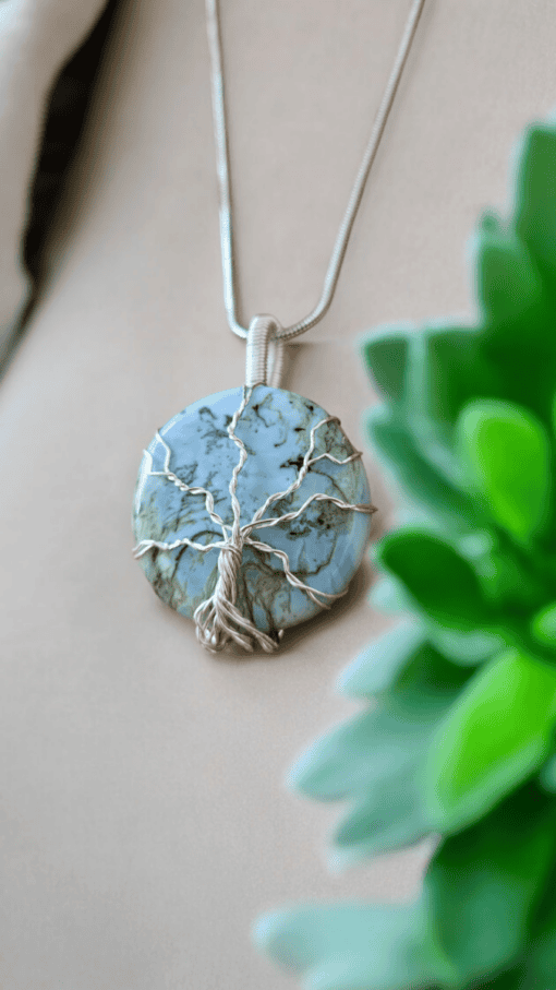 Silver wire-wrapped blue stone necklace with a tree of life design, displayed near a green plant on a beige surface.