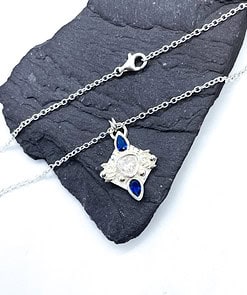 Delicate silver necklace with a pendant featuring a central clear gemstone and two blue teardrop stones on a dark slate.