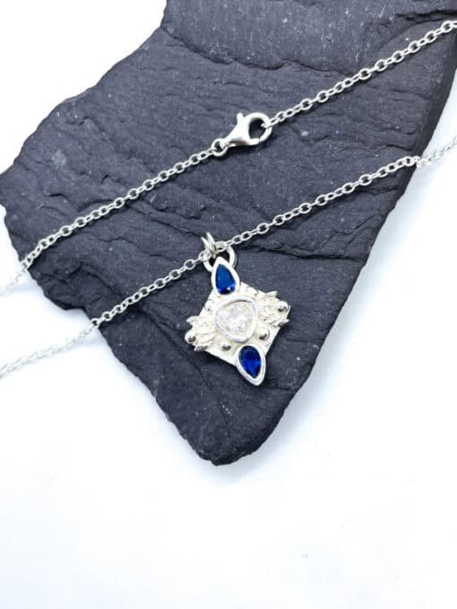 Delicate silver necklace with a pendant featuring a central clear gemstone and two blue teardrop stones on a dark slate.