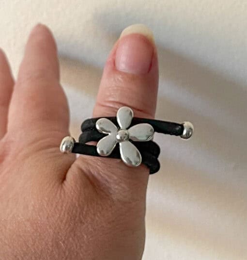 Silver flower ring with a black band worn on a thumb against a neutral background.