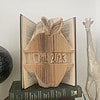 A folded book art piece showing an apple shape with "Oct 2023," surrounded by hardcover books, a globe, and a giraffe statue.