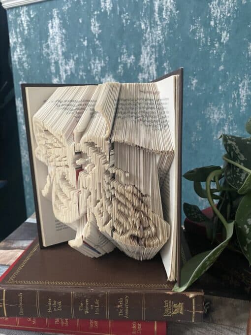 An open book with intricate folded pages forming a pattern, placed on top of other books near a potted plant.