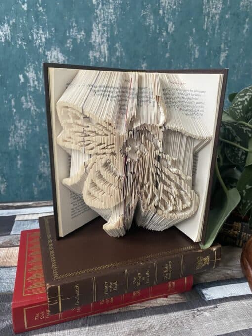 Open book with intricately folded pages forming a butterfly design, placed on top of two closed books on a wooden surface.