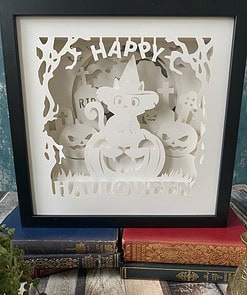 Framed paper art of a Halloween scene with a cat in a witch hat, pumpkins, and gravestones, placed on stacked vintage books.