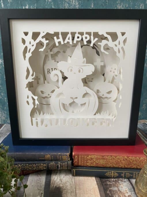 Framed paper art of a Halloween scene with a cat in a witch hat, pumpkins, and gravestones, placed on stacked vintage books.