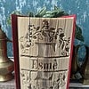 Opened book with intricately folded pages creating a 3D design, displayed on a table with brass artifacts and green plant in background.