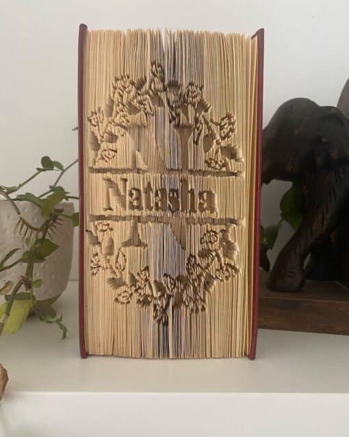 Folded book art displaying intricate tree designs around the word "Natasha," with plants and a wooden elephant in the background.