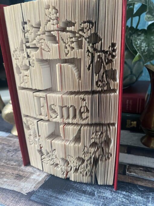 A book with intricately folded pages creating a 3D image and "Family" text, positioned on a wooden table with leafy plants in the background.